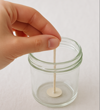 A hand pressing a wick sticker into a glass jar, with the wick standing upright